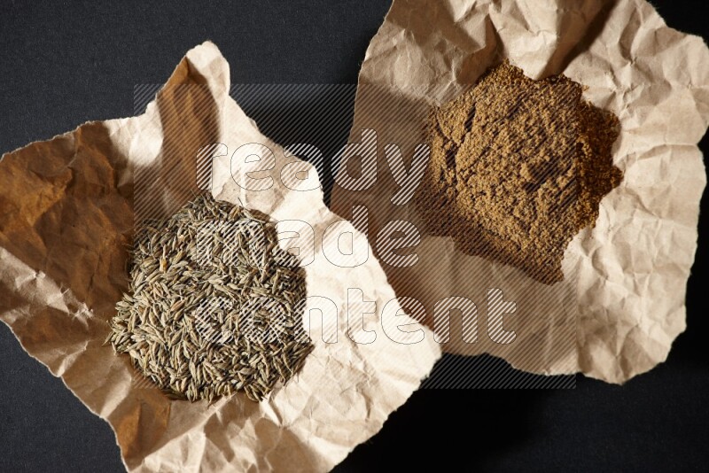 Cumin seeds and powder in 2 crumpled pieces of paper on black flooring