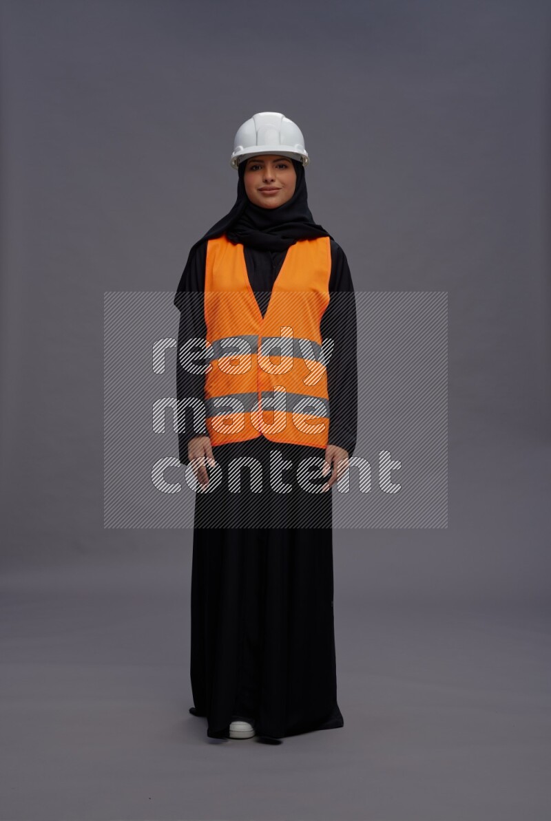 Saudi woman wearing Abaya with engineer vest standing interacting with the camera on gray background