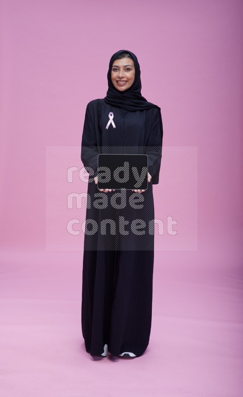 Saudi woman wearing pink ribbon on Abaya standing showing a tablet to camera on pink background