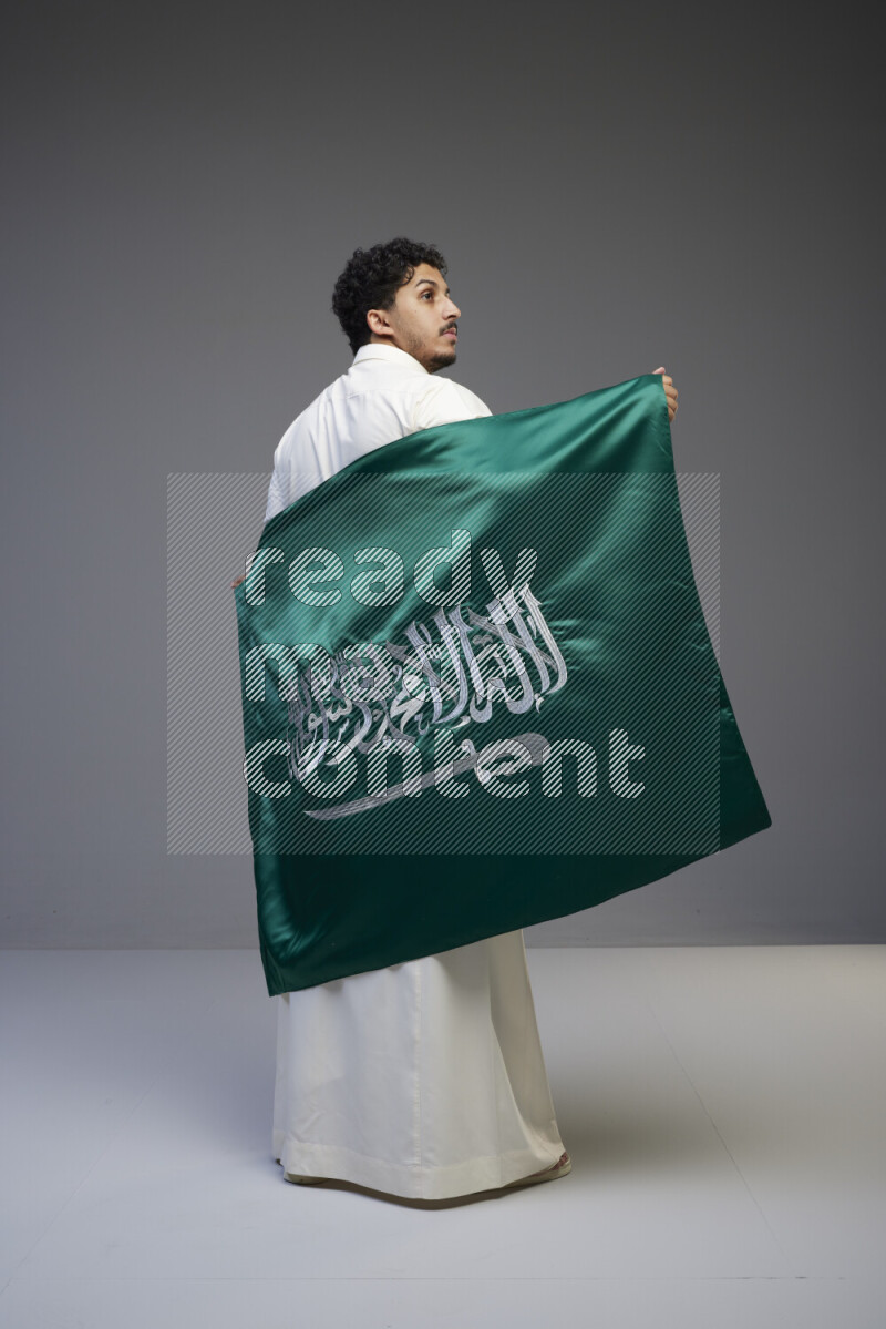 A saudi man standing wearing thob holding big saudi flag on gray background