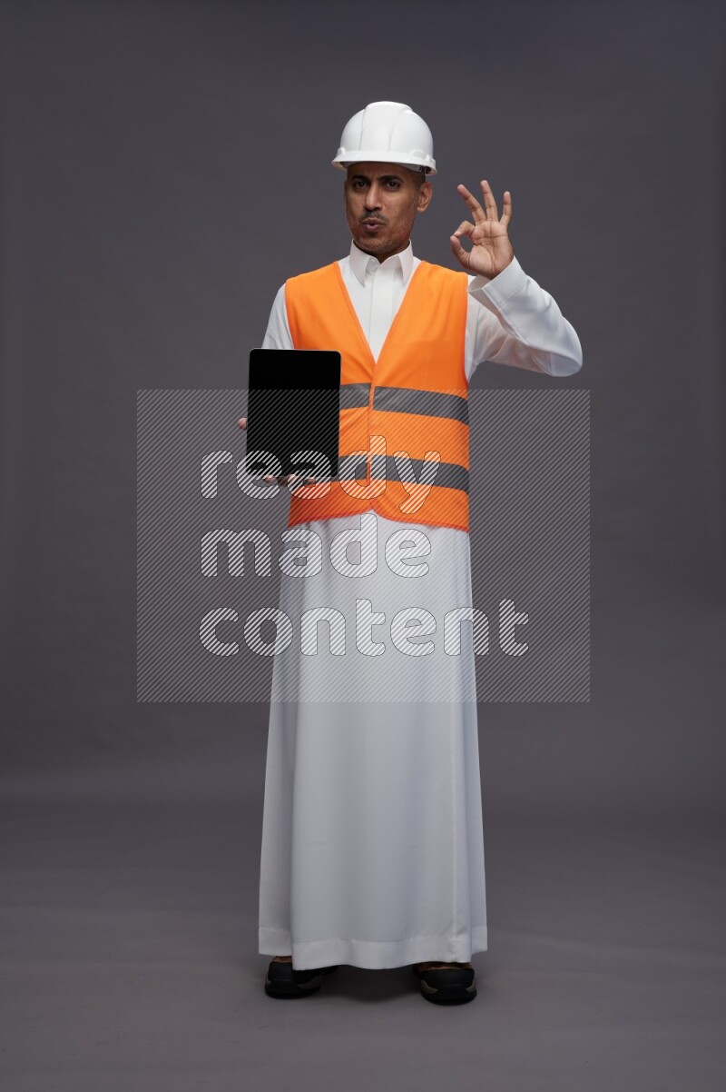 Saudi man wearing thob with engineer vest standing showing tablet to camera on gray background