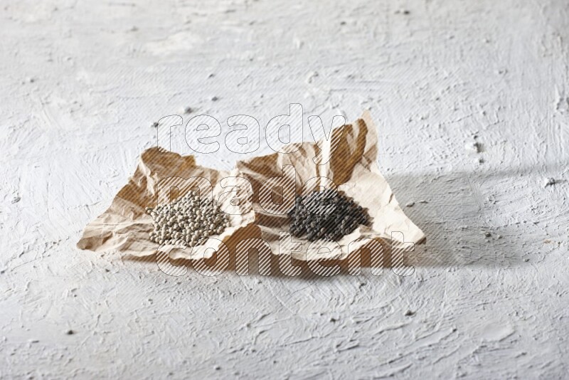 2 Crumpled pieces of paper full of black and white pepper beads on a textured white flooring