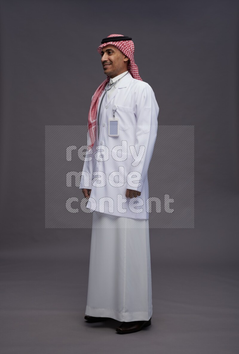 Saudi man wearing thob with lab coat and shomag with pocket employee badge with stethoscope standing interacting with the camera on gray background