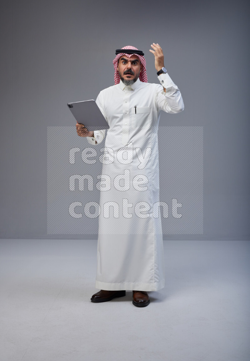 Saudi man Wearing Thob and red Shomag standing working on tablet on Gray background