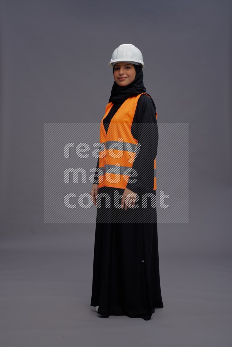 Saudi woman wearing Abaya with engineer vest standing interacting with the camera on gray background