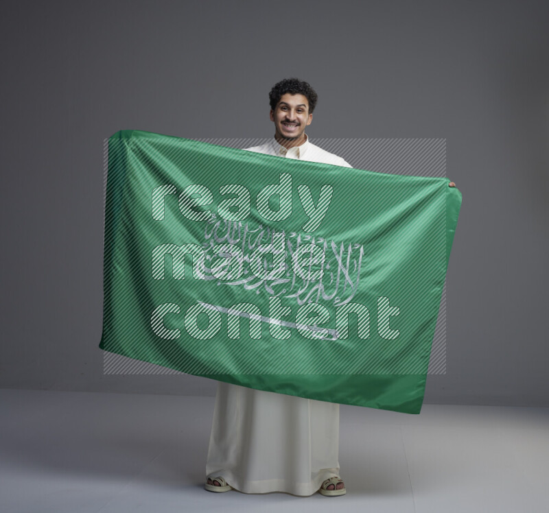 A Saudi man standing wearing thob holding big Saudi flag on gray background