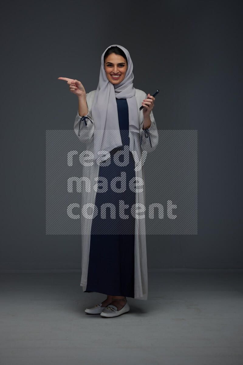 A Saudi woman wearing a light gray Abaya and head scarf standing and holding a phone on a grey background