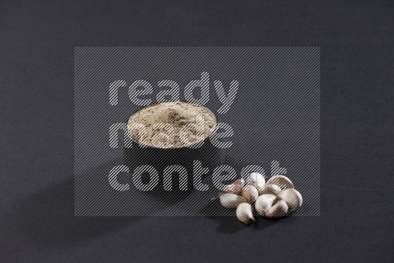 A black pottery bowl full of garlic powder with some cloves beside it on a black flooring