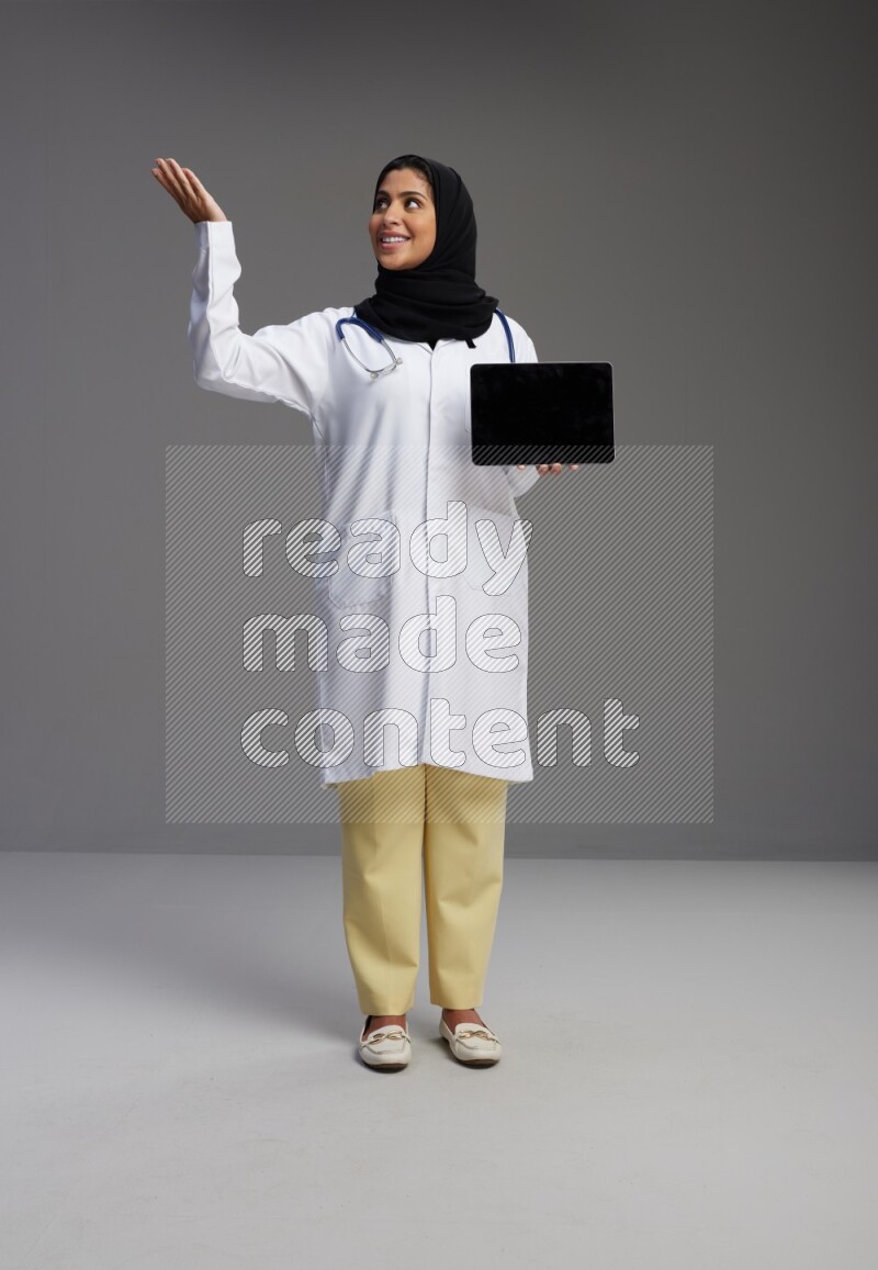Saudi woman wearing lab coat with stethoscope standing showing tablet to camera with sign in the back on Gray background