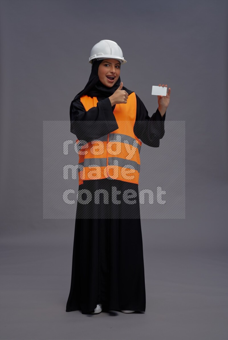 Saudi woman wearing Abaya with engineer vest standing holding ATM card on gray background