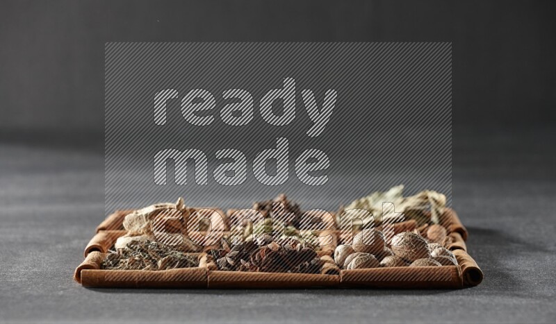 9 squares of cinnamon sticks full of cardamom in the middle surrounded by nutmeg, cinnamon, bay laurel leaves, cloves, cumin, dried ginger, dried basil and star anise on black flooring