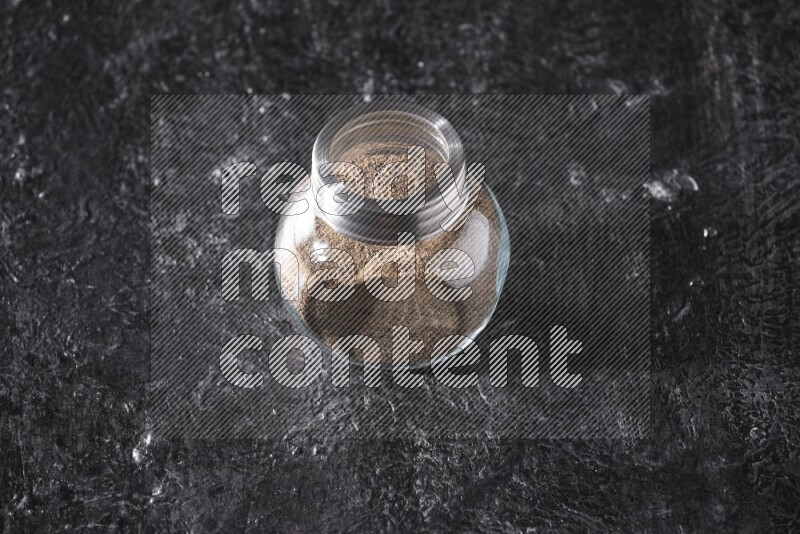A glass spice jar full of black pepper powder on textured black flooring
