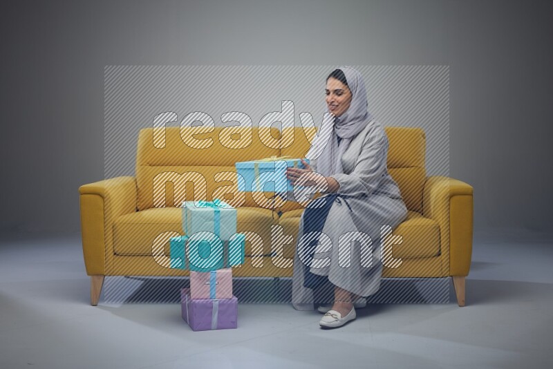 A Saudi woman wearing a light gray Abaya and head scarf sitting on a yellow sofa holding a gift box beside multi color gift boxes eye level on a grey background