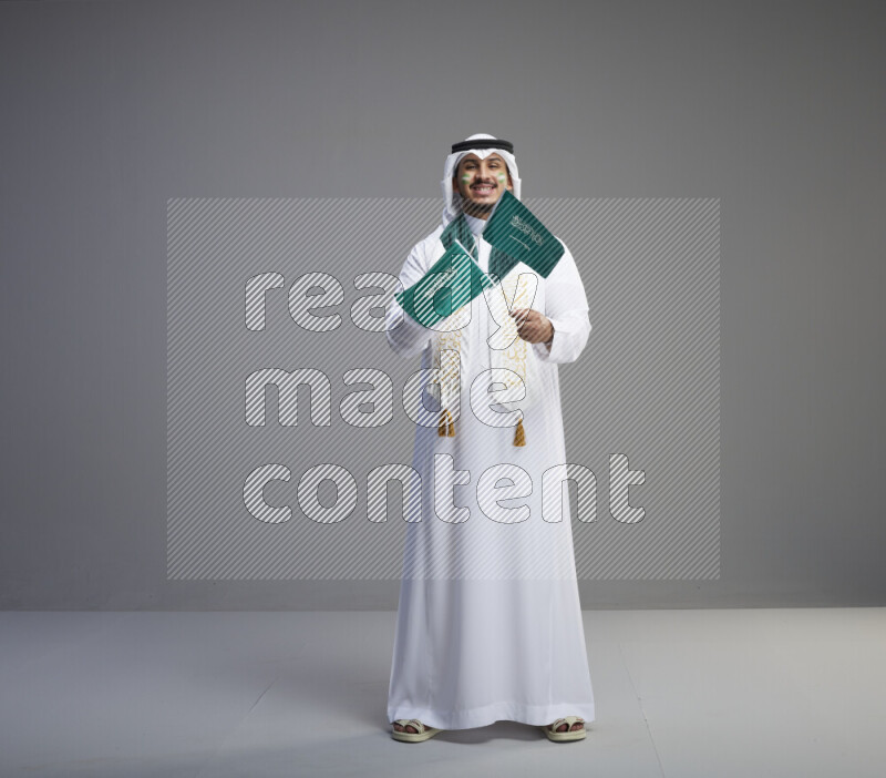 A saudi man standing wearing thob and white shomag with face painting and saudi flag scarf and holding small saudi flag on gray background