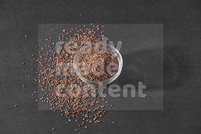 A glass bowl full of flaxseeds surrounded by the seeds on a black flooring