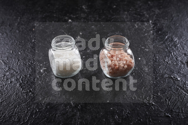 2 glass jars one is filled with coarse himalayan salt and the other with coarse sea salt on black background