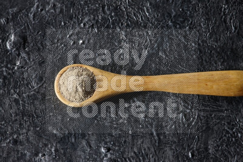A wooden spoon full of white pepper powder on textured black flooring