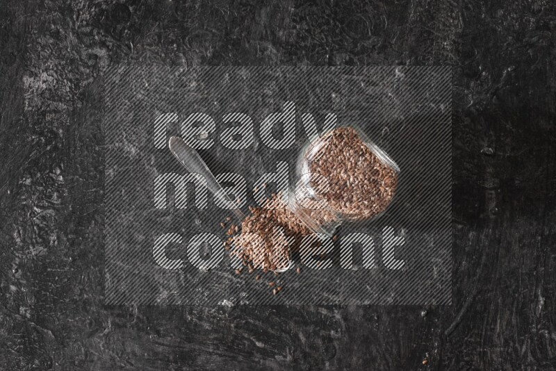 A glass spice jar full of flaxseeds flipped and seeds spread out with a metal spoon full of the seeds on a textured black flooring