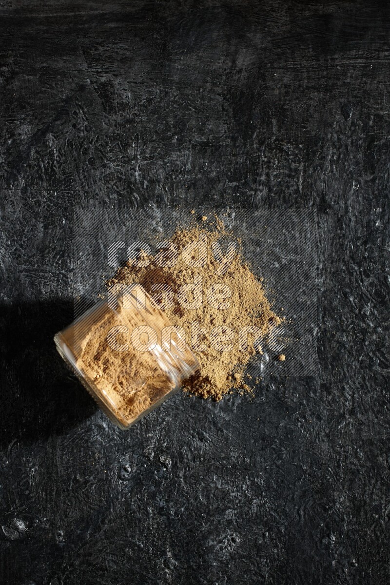 A flipped glass jar full of allspice powder and powder spilled out of it on a textured black flooring