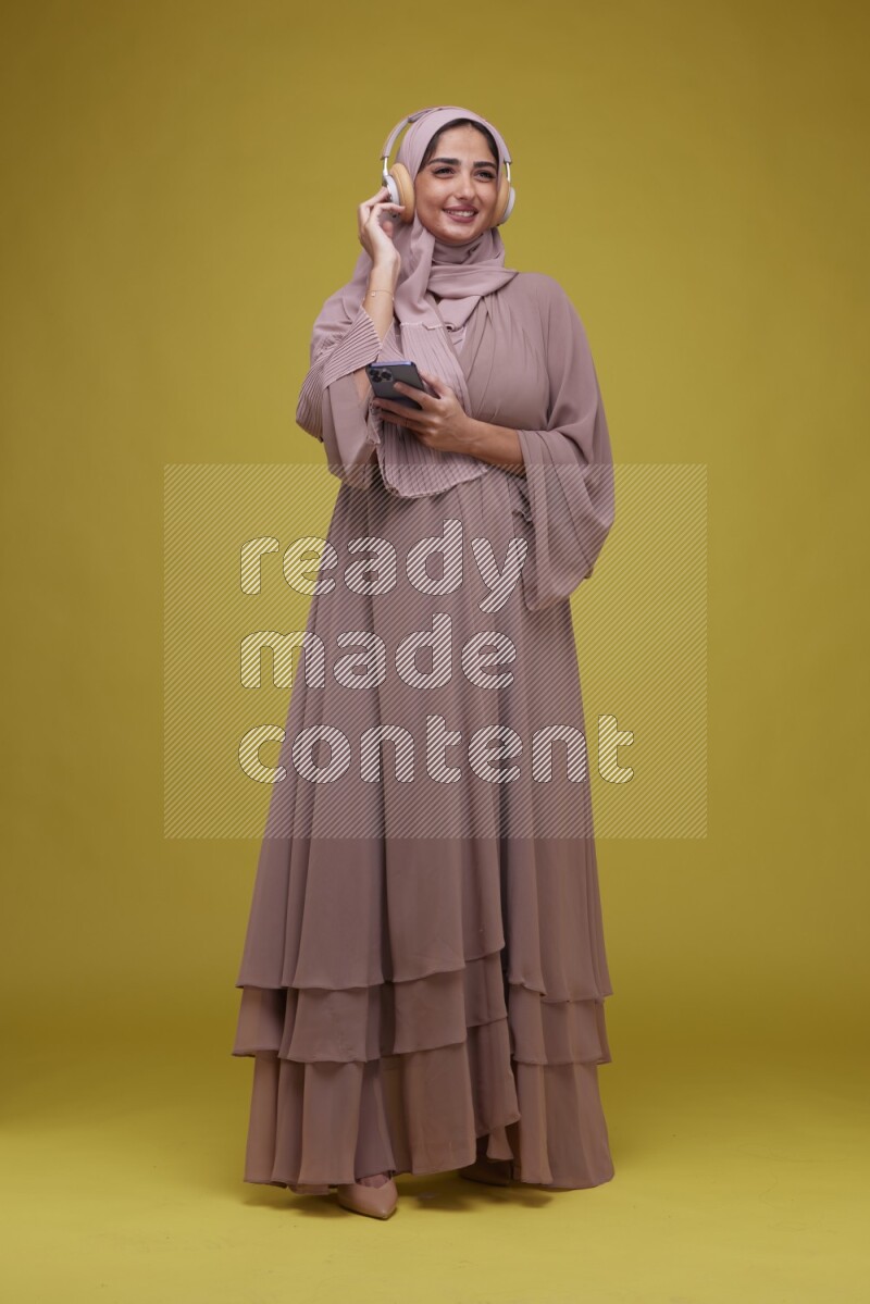 A woman Listening to Music on a Yellow Background wearing Brown Abaya with Hijab