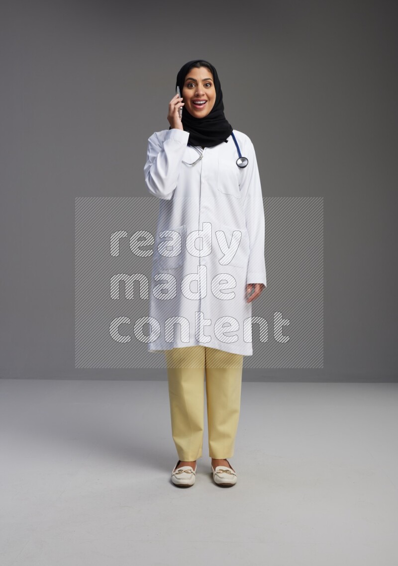 Saudi woman wearing lab coat with stethoscope standing talking on phone on Gray background
