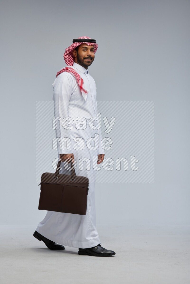 Saudi man Wearing Thob and shomag standing holding bag on Gray background