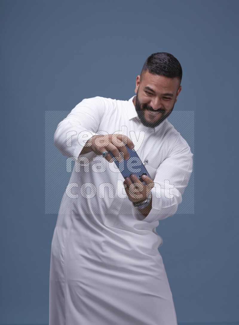 A man Playing a Game on his Smartphone  on Blue Background wearing Saudi Thob