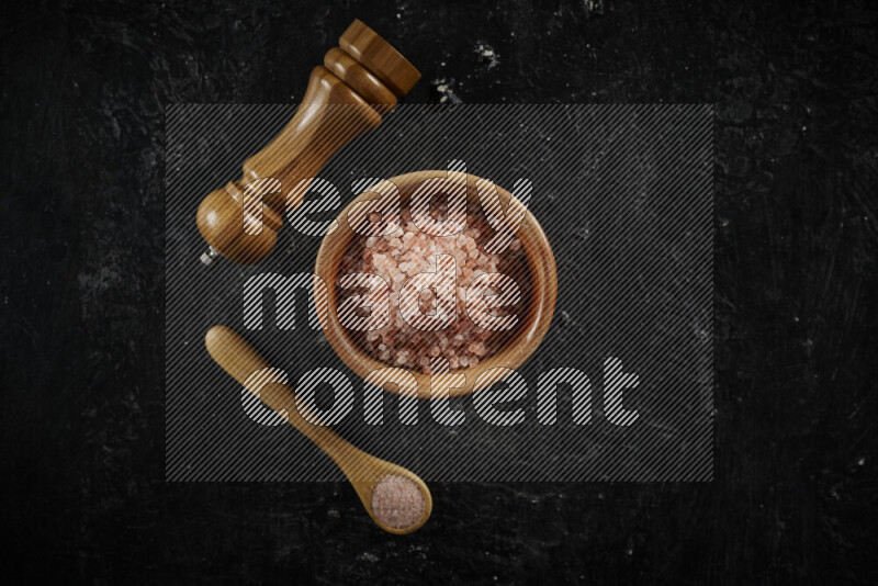A wooden bowl and spoon filled with coarse pink himalayan salt and a wooden grinder beside them on black background