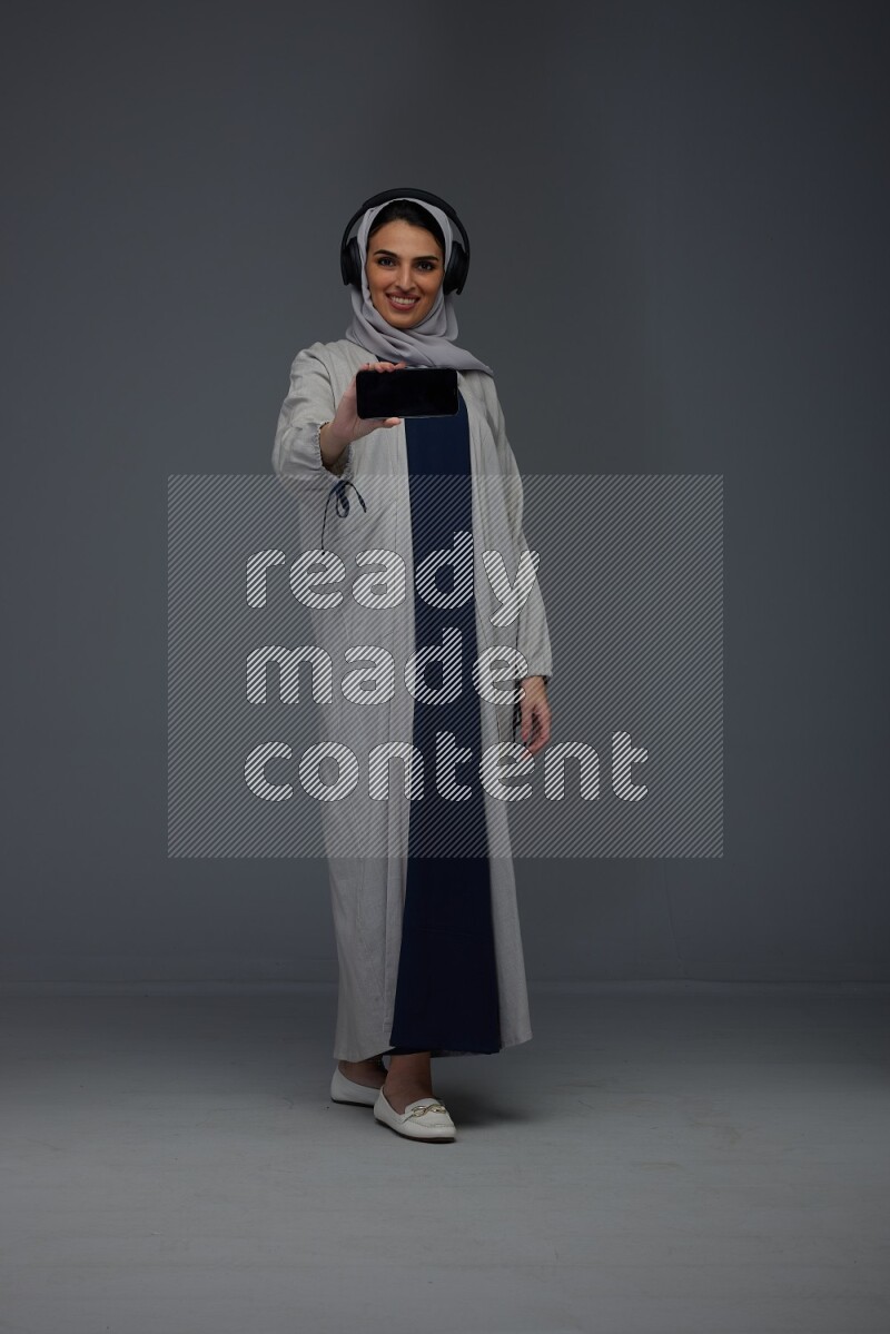 A Saudi woman wearing a light gray Abaya and head scarf setting on a dark grey chair and watching on the tablet eye level on a grey background
