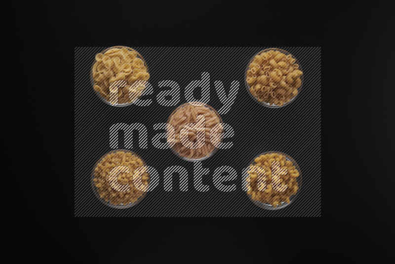 Different pasta types in 5 glass bowls on black background