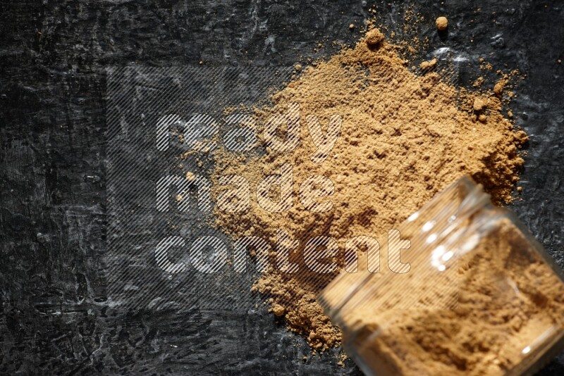 A flipped glass jar full of allspice powder and powder spilled out of it on a textured black flooring