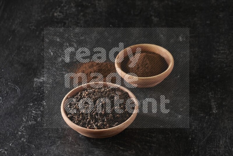 2 wooden bowls full of cloves powder and whole cloves on a textured black flooring