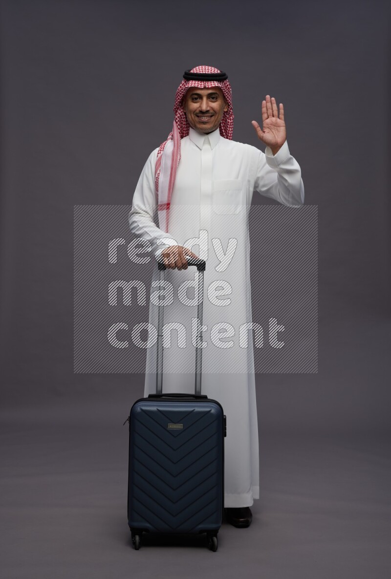 Saudi man wearing thob and shomag standing holding bag on gray background