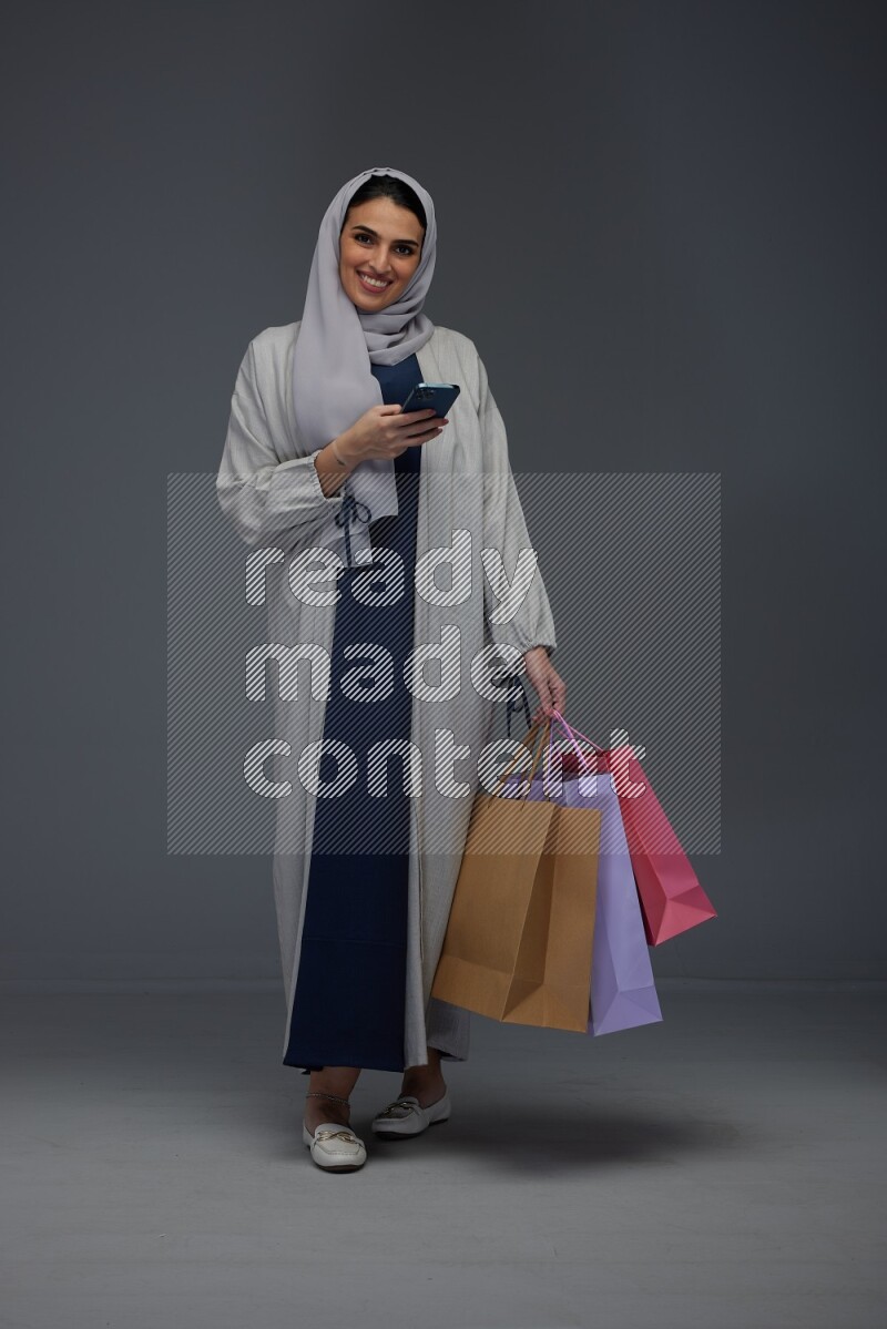 A Saudi woman wearing a light gray Abaya and head scarf standing and holding shopping bags making different poses eye level on a grey background