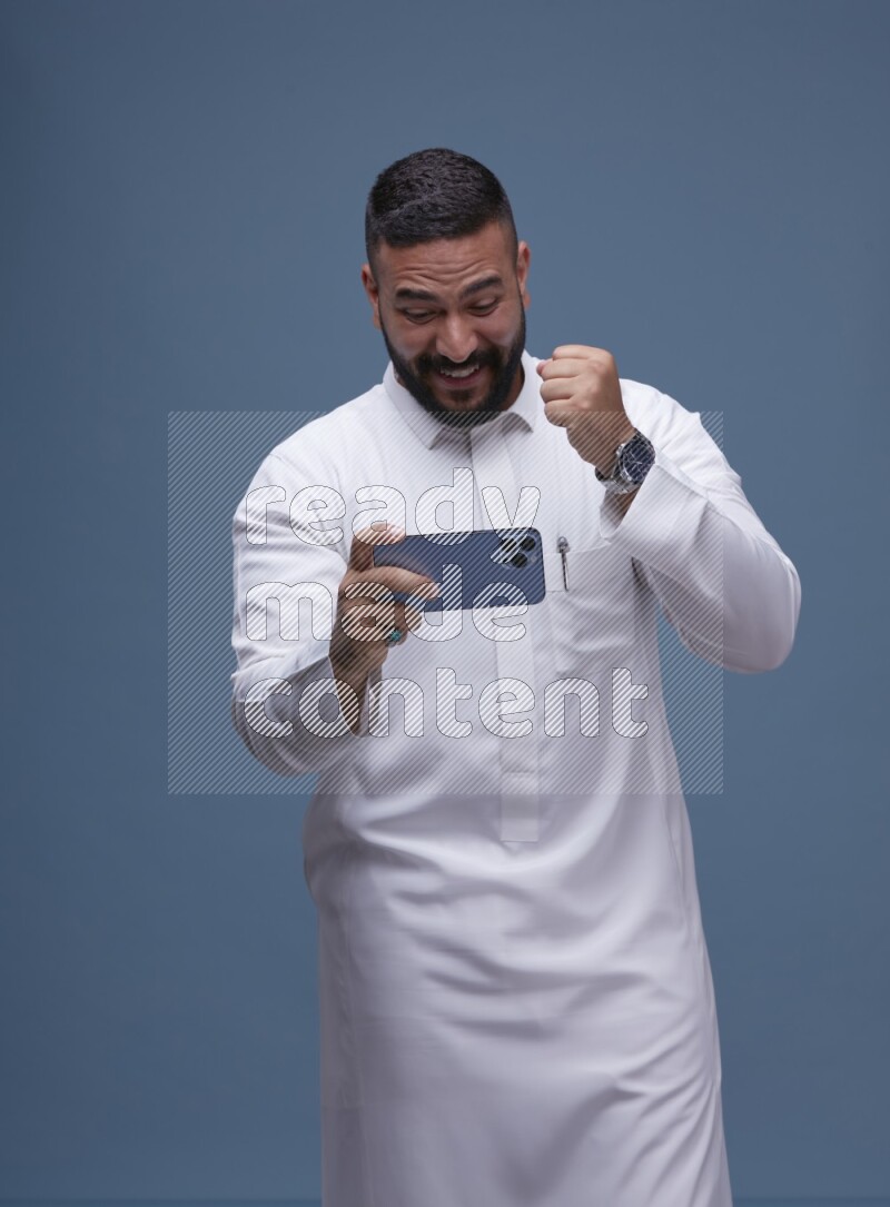 A man Playing a Game on his Smartphone  on Blue Background wearing Saudi Thob