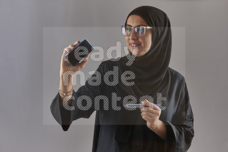 Woman in black abaya, hijab and glasses holding a marker pen to write on a transparent board with different reactions and expressions on grey background