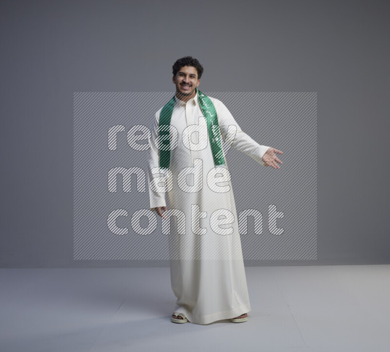 A Saudi man standing wearing thob and Saudi flag scarf on gray background