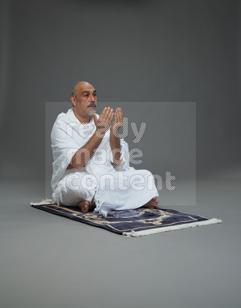 A man wearing Ehram sitting on prayer mat dua'a on gray background