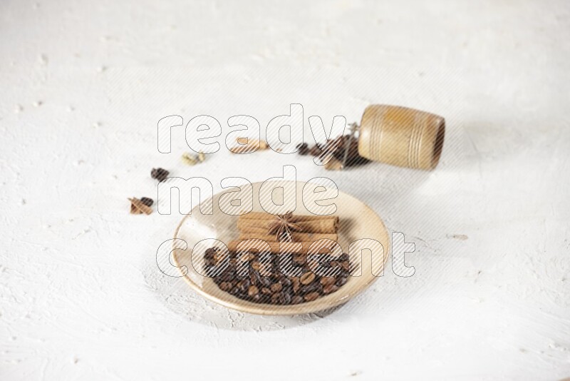 Beige plate full of coffee beans, cinnamon sticks and star anise with a coffee grinder, coffee beans, cinnamon pieces and cardamom next of it on white background