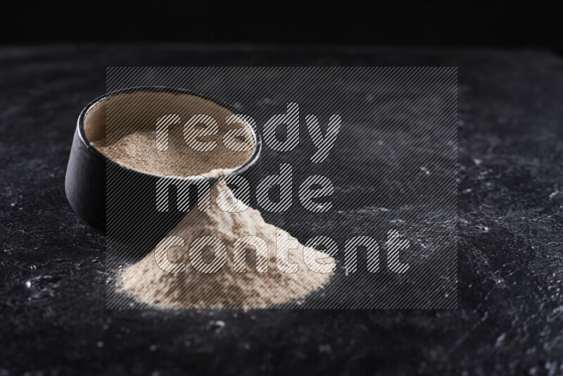 A black pottery bowl full of onion powder with fallen powder from it on black background