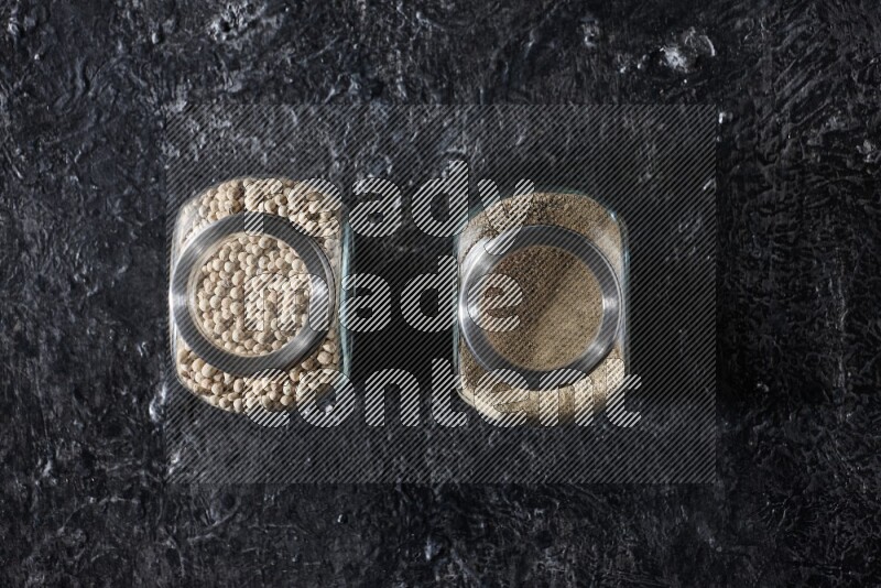 2 Herbal glass jars full of white pepper beads and powder on textured black flooring