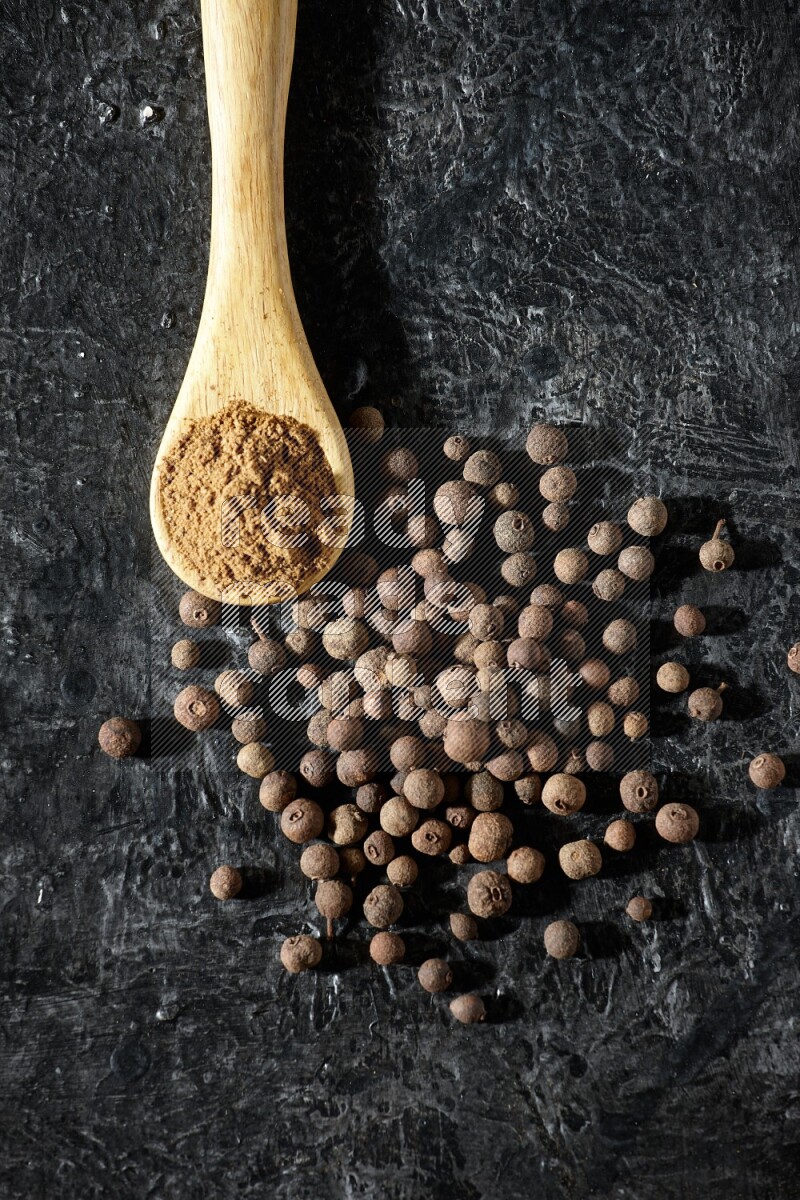 A wooden spoon full of allspice powder and whole balls spreaded on a textured black flooring