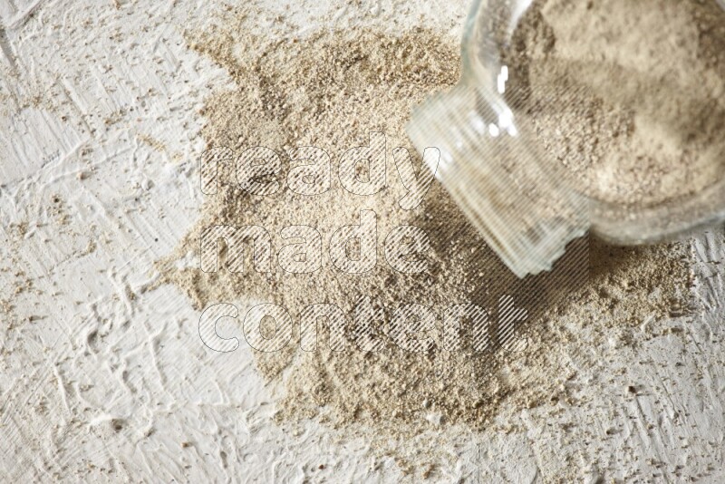 A flipped herbal glass jar full of white pepper powder with spilled powder on textured white flooring