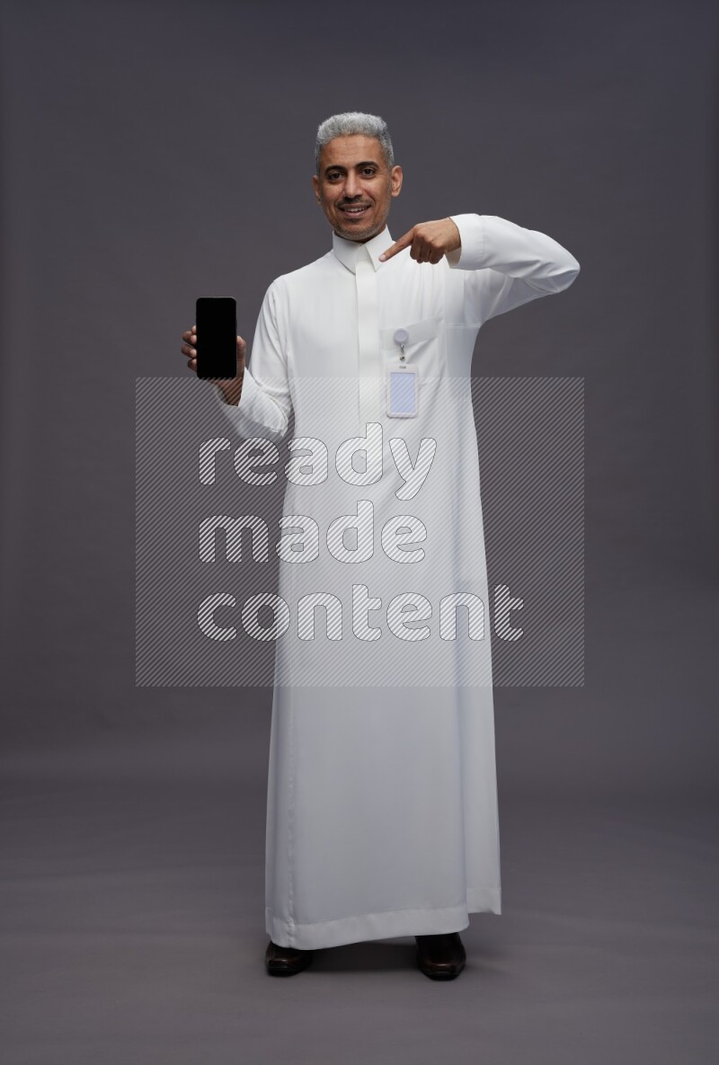 Saudi man wearing thob with pocket employee badge standing showing phone to camera on gray background