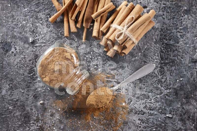 Herbal glass jar full cinnamon powder flipped and a metal spoon full of powder, cinnamon sticks stacked and bounded in the back on textured black background in different angles