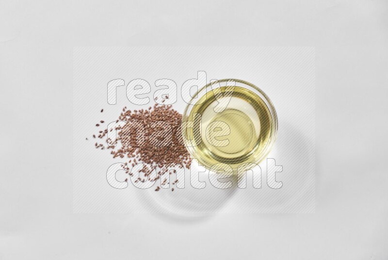 A glass bowl full of flaxseeds oil and flaxseeds beside it on a white flooring