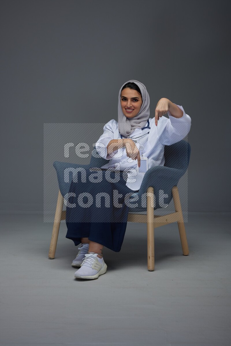 A doctor wearing a light gray head scarf setting on a dark grey chair and using a phone eye level on a grey background