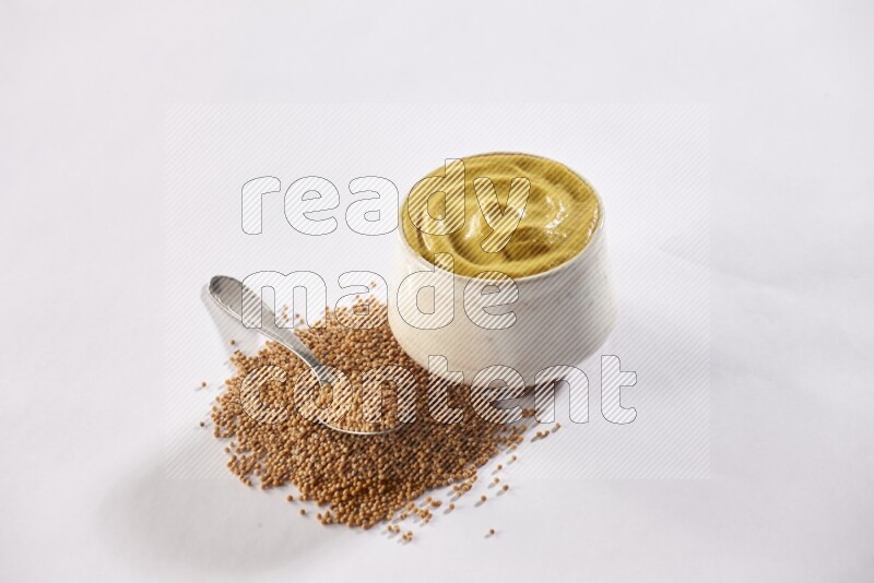 A beige pottery bowl full of mustard paste with mustard seeds underneath with a metal spoon on white flooring