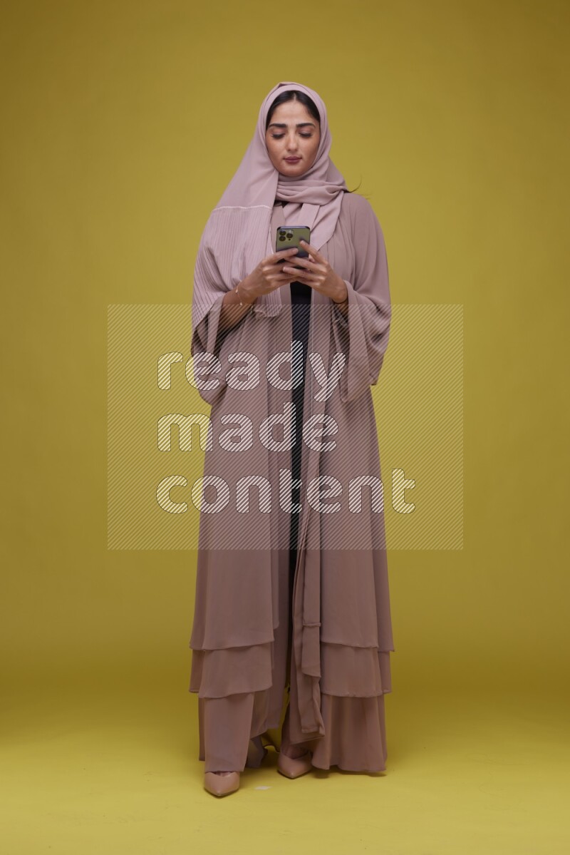 A woman Texting on her phone on a Yellow Background wearing Brown Abaya with Hijab