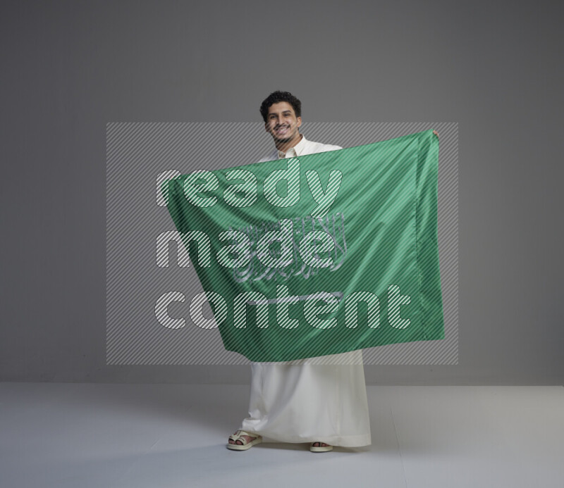 A Saudi man standing wearing thob holding big Saudi flag on gray background